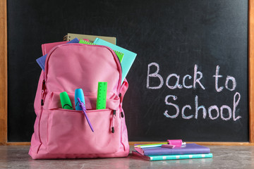 Backpack with stationery on table against blackboard with written words BACK TO SCHOOL