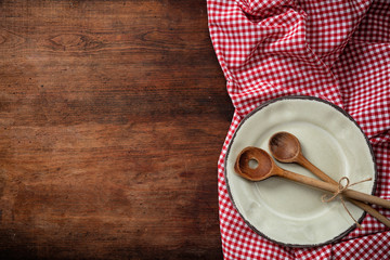 Empty plate, kitchen utensils and red tablecloth on wooden table, top view, copy space