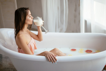 Wellness and relaxation concept. Half-naked slender woman holds a glass of milk relaxes in white milky bath, takes care of skin and appearance. Female recreats in spa center