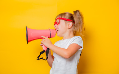 Portrait of an emotional toddler girl in glasses with megaphone on yellow background.