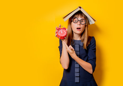 Portrait of a nerd girl in glasses with books and alarm clock on yellow background