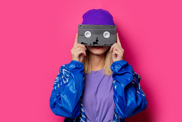 Young blonde girl in 90s sports jacket and VHS cassette on pink background.