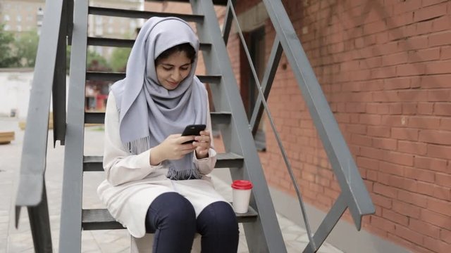 Attractive young woman with black hair wearing traditional muslim clothes smiling, sitting on iron stairs in a city and texting. Slider real time establishing shot