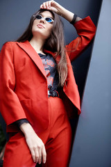 Fashion young girl posing. Stylish woman in a red suit and sunglasses . Against grey background