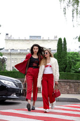 Two fashion young girl walk across the road. Stylish woman in a red suit and sunglasses and in white fur and sport red pants with .Emotioanl shot.Motion.