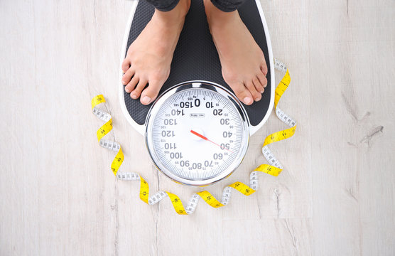 Woman measuring her weight using scales near tape on floor, top view