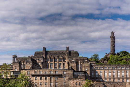 Edinburgh, Scotland, UK - June 13, 2012: Large Brown Building Was Old Royal High School And Abandoned Site Of New Scottish Assembly With Nelson Monument In Back, All Under Blue Cloudy Sky.