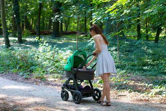 Forest walk with stroller, side view of young mom in beautiful dress walking on the pathway with her infant in the pram, enjoying fresh air and healing after postpartum, mother and baby stroller walk