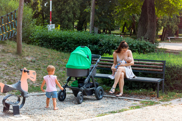 Motherhood, young pretty mother on parental leave breastfeeding her baby in children playground,...