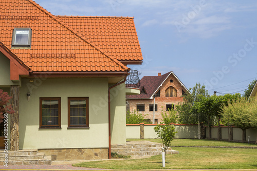 New Nice One Story Cottage With Shingle Roof Balcony Plastic Attic New Nice One Story Cottage With Shingle Roof Balcony Plastic Attic Windows Paved Yard Green Lawn And Decorative Blooming Trees On Bright Blue Sky