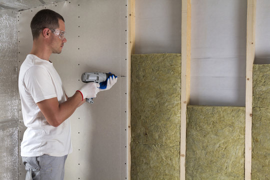 Worker in goggles with screwdriver working on insulation. Drywall on wall beams, insulating rock wool staff in wooden frame. Comfortable warm home, economy, construction and renovation concept.