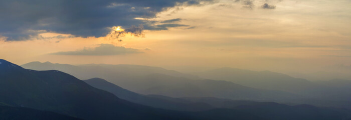 Wide panorama, fantastic view of covered with morning mist green Carpathian mountains at dawn under dark clouds and light pink sky before sunrise. Beauty of nature, tourism and traveling concept.
