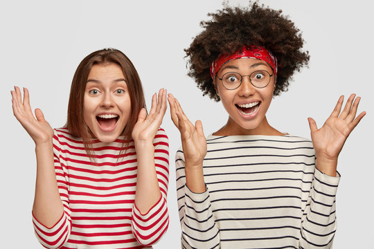 Horizontal shot of beautiful amazed females clasp hands with happiness, meet with their best friend, dressed casually, isolated over white background. People, race and positive emotions concept