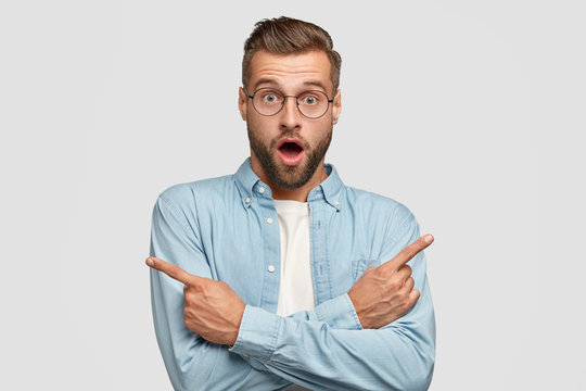 Shocked Caucasian male points at different sides with index fingers, cant choose between two items, has puzzled expression, wears round glasses and blue shirt, isolated over white background