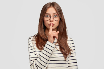 Adorable young woman in casual clothes, makes silence gesture, looks directly at camera, touches lips with index finger, tries to be secret, isolated on white background. People and conspiracy concept