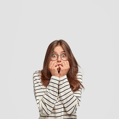 Frightened beautiful young woman bites finger nails, looks anxiously at camera, dressed in fashionable clothes, has appealing look, stands against white background. People, emotions concept.