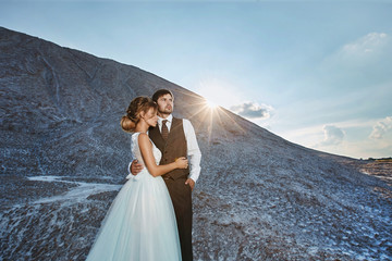 Fashionable and beautiful couple, sexy and elegant blonde model girl with stylish hairstyle, in white lace dress and stylish handsome men in trendy suit posing outdoors at the sunset at the mountains