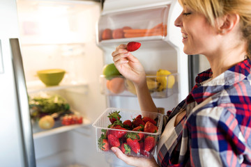 Pretty young woman picking strawberries of fridge in the kitchen.