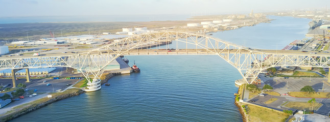 Panorama aerial view of Corpus Christi Harbor Bridge with row of oil tanks and wind turbines farm in distance. A through arch bridge crosses the Corpus Christi Ship Channel