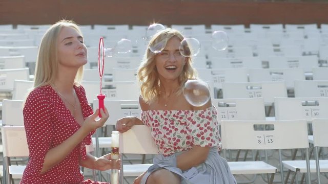 Two attractive beautiful young blond women blow bubbles in the park and are happy on sunset. Girlfriends in park. Slow motion 
