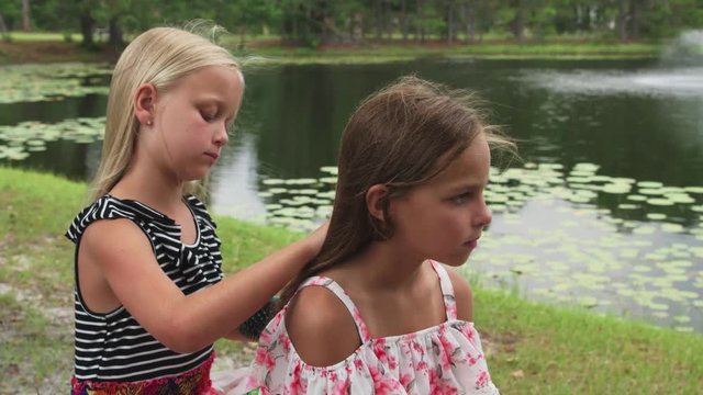 Two young best girl friends spend time together outdoors by lake brushing hair.