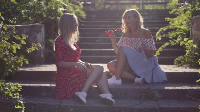 Two attractive beautiful young blond women blow bubbles in the park and are happy on sunset. Girlfriends in park on the steps