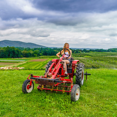 Obraz premium Beautiful woman on a red agricultural tractor, green field, landscape of meadow with grass Upstate New York
