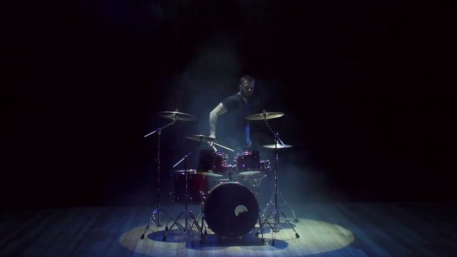 A male drummer plays a drum set on stage in the dark, he finishes the performance gets up and leaves the stage. Slow motion.