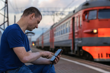 A man is sitting at the train station of electric trains and what he is looking for in a smartphone