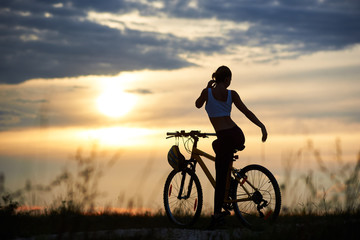 Fototapeta na wymiar Silhouette of sporty female cyclist, wearing sportswear and posing near her bicycle on trail. Slender incognito woman enjoying nature and observing wonderful landscapes and amazing sunset.