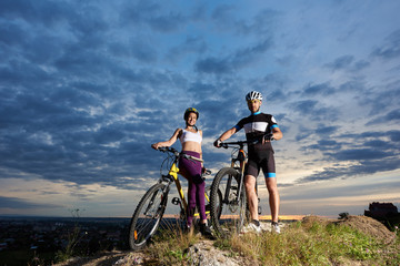 Fototapeta na wymiar Two positive cyclists holding hands on handle bars of their bikes, looking at camera and smiling. Beautiful girl wearing violet leggings and white top, man in black sportswear and sport glasses.
