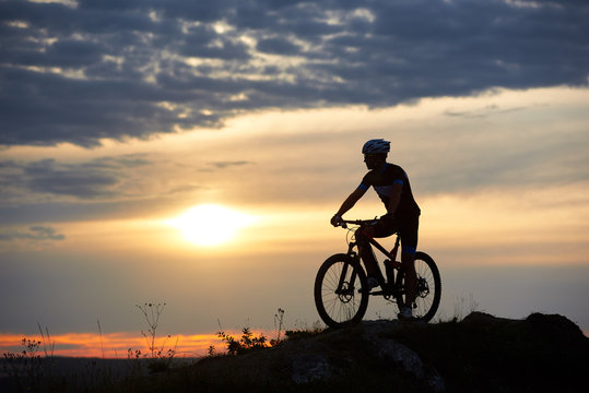 Robust and strong man wearing sportswear and helmet sitting on his bike and posing on hill. Cyclist enjoying great view of sunset. Concept of motivation, recreation and healthy activities.