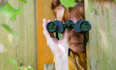 eine neugierige Frau blickt mit einem Fernglas durch einen Zaun - obrazy, fototapety, plakaty