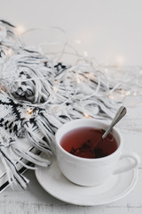 Cup of red hibiscus tea, book, lights for home decoration and woolen knitted blanket on wooden table, vertical photo. Hygge concept