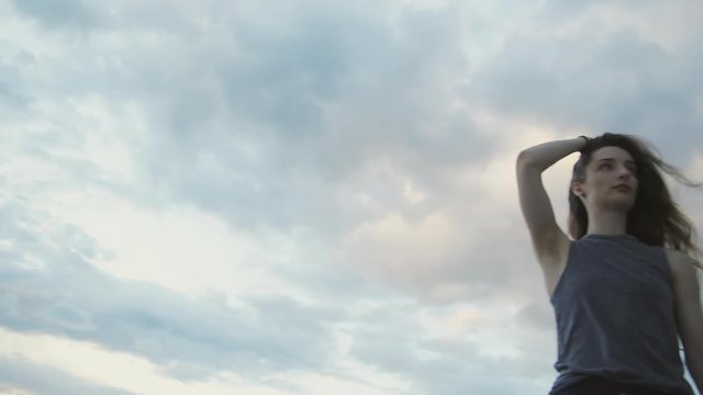Camera pans from the sky, a girl is standing in front of a late evening blue sky, waving her hair