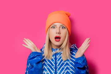 Young blonde girl in 90s sports jacket and hat on pink background.