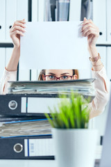 Obraz premium Young female office worker hides behind the folder holding the clean white sheet of paper. Concept of low pay, long working day.