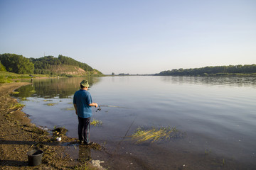 Fototapeta na wymiar a man on a summer clear day, in a blue T-shirt with a fishing rod, stands on the river bank and catches fish