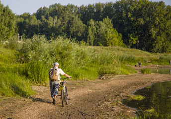 an elderly fisherman man walks along the river bank and pushes a bicycle