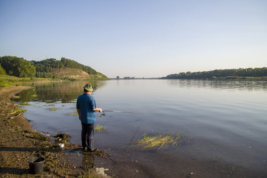 a man on a summer clear day, in a blue T-shirt with a fishing rod, stands on the river bank and catches fish