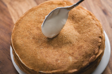 Adiing sour cream to stack of fresh hot crepes or blinis on wood table