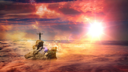 A man with spread arms standing on a rock protruding above the clouds and looking at the rising sun.