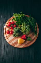 Fresh vegetables: cherry tomatoes, broccoli, pepper, dill, parsley on a round wooden board, on a dark wooden table. Recipe. Ingredients. Dietary food. Place under the text. View from above