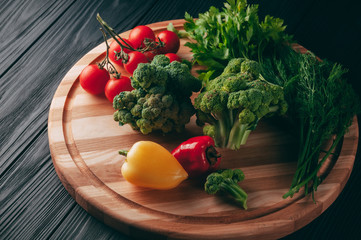 Fresh vegetables: cherry tomatoes, broccoli, pepper, dill, parsley on a round wooden board, on a dark wooden table. Recipe. Ingredients. Dietary food. Place under the text. View from above