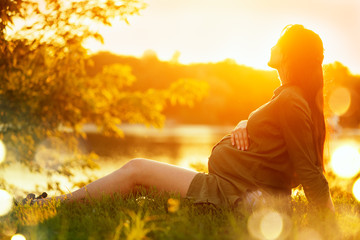 Pregnant woman sitting on green grass in summer park, enjoying nature. Healthy pregnancy concept