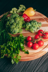 Fresh vegetables: cherry tomatoes, broccoli, pepper, dill, parsley on a round wooden board, on a dark wooden table. Recipe. Ingredients. Dietary food. Place under the text. View from above