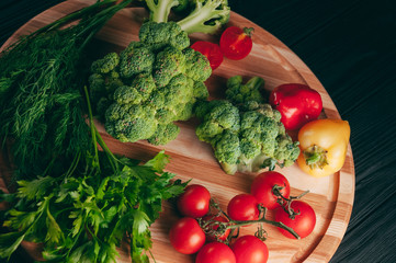 Fresh vegetables: cherry tomatoes, broccoli, pepper, dill, parsley on a round wooden board, on a dark wooden table. Recipe. Ingredients. Dietary food. Place under the text. View from above
