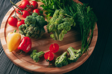 Fresh vegetables: cherry tomatoes, broccoli, pepper, dill, parsley on a round wooden board, on a dark wooden table. Recipe. Ingredients. Dietary food. Place under the text. View from above