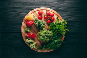 Fresh vegetables: cherry tomatoes, broccoli, pepper, dill, parsley on a round wooden board, on a dark wooden table. Recipe. Ingredients. Dietary food. Place under the text. View from above