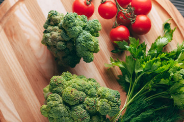 On a dark wooden table, on a round wooden board fresh green broccoli, parsley, dill and cherry tomatoes for your health. Recipe. Ingredients. Dietary food. Place under the text. View from above